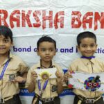 Three young boys in beige uniforms display handmade rakhi bracelets in front of a raksha bandhan banner